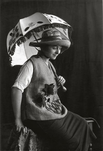 Una mujer sentada y vestida con sombrero, sombrilla, chaleco y falda.