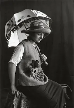 Una mujer sentada y vestida con sombrero, sombrilla, chaleco y falda.