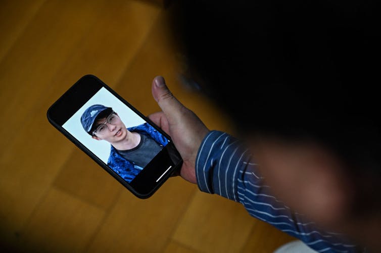 A person holding a phone with the face of a young man wearing a baseball cap on its screen.
