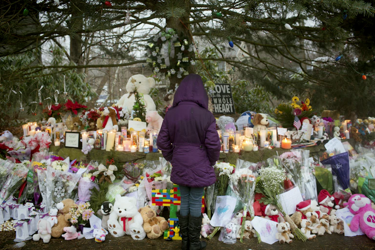A woman in a puffy purple jacket is seen from behind, contemplating a large spontaneous memorial to the many children and educators killed in the Sandy Hook Elementary School shooting.
