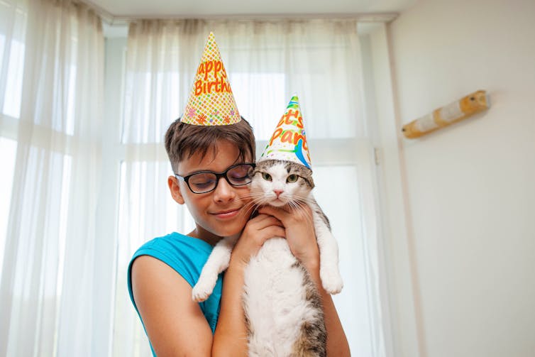 A boy holding up a cat, both wearing birthday hats.
