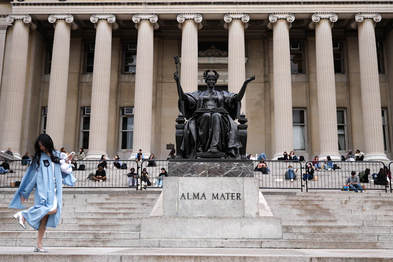 A large statue of a person seated sits in front of a large building with columns. People stand and sit on the steps in front of the building.