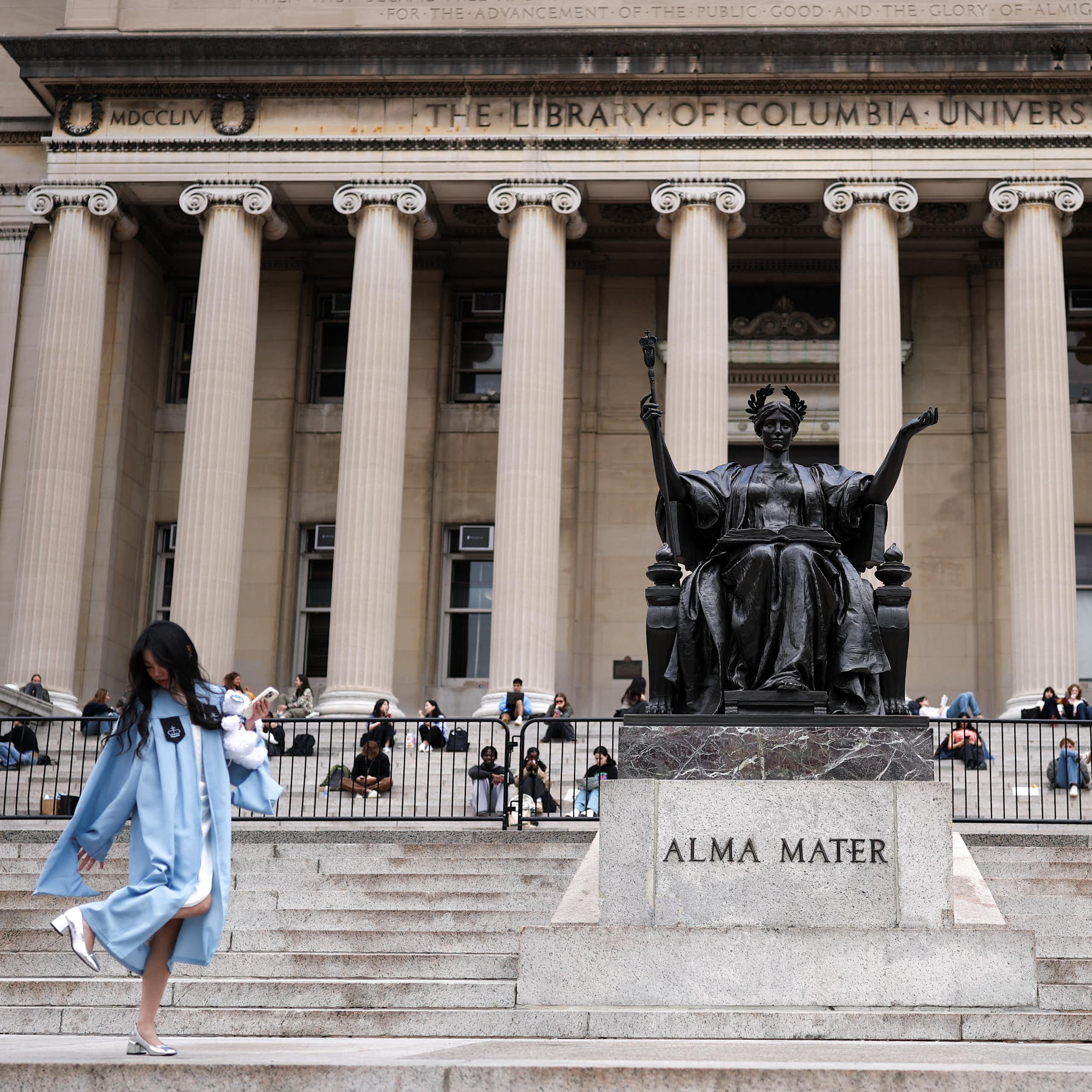 A large statue of a person seated sits in front of a large building with columns. People stand and sit on the steps in front of the building.