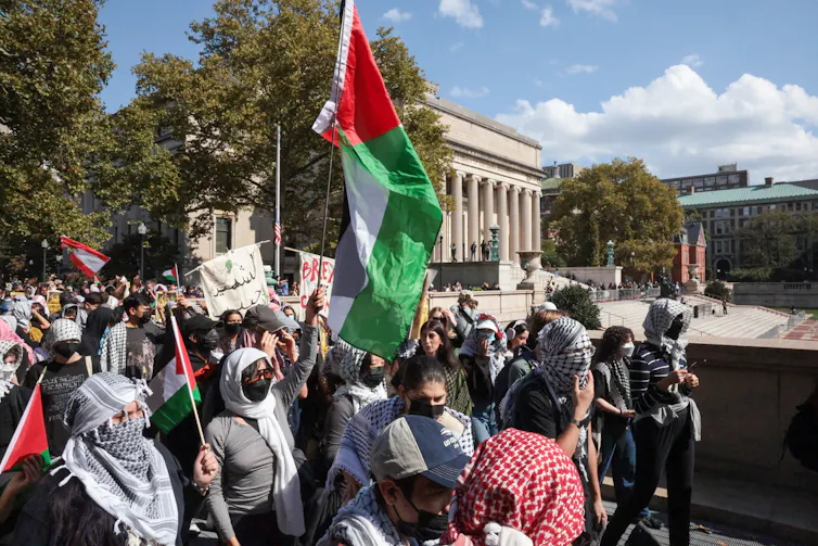 A group of people with their faces covered by cloth hold red, green, white and black flags and walk together in front of a large building with columns.