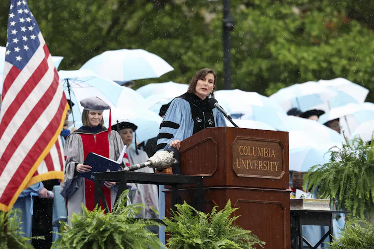 A woman wears a blue robe and stands at a wooden podium. People dressed in caps and gowns for graduation stand behind her and hold light blue umbrellas.