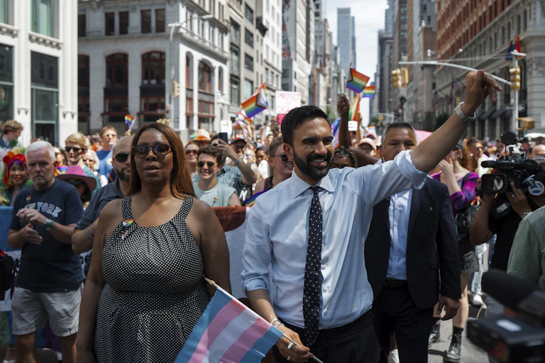 A man walking in a parade waves to spectators as hundreds of people walk behind him.