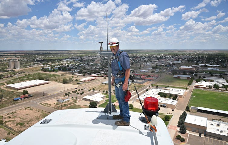 A worker wearing a hardhat stands on top of a wind turbine, with a wide view of the landscape around him.