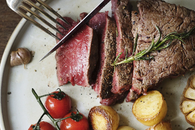 Knife and fork used to cut rare steak on plate with cherry tomatoes and potatoes