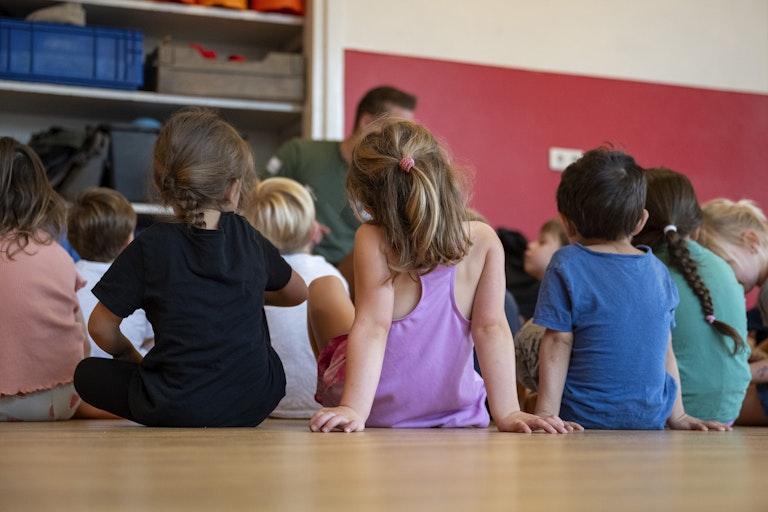 A group of small children sit on the floor around a teacher.