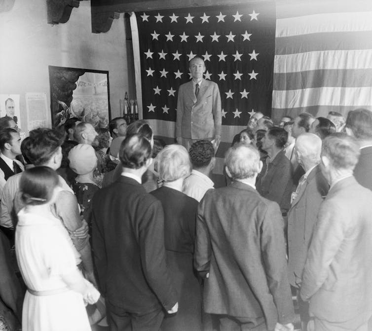 A black and white photo shows a man on a stage, the American flag behind him, speaking to a crowd.
