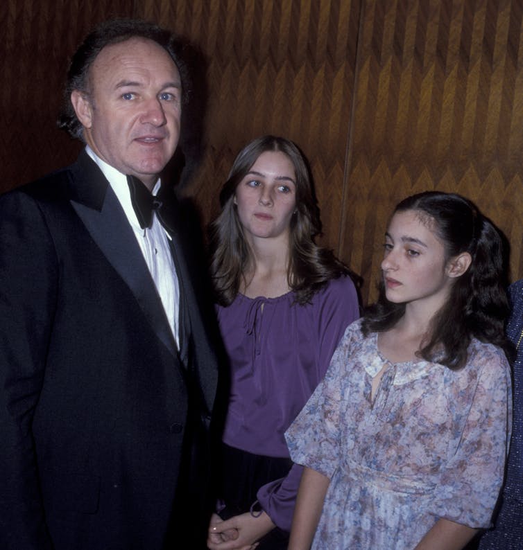 Man in tuxedo and a large bowtie stands next to two teenagers who are looking away.