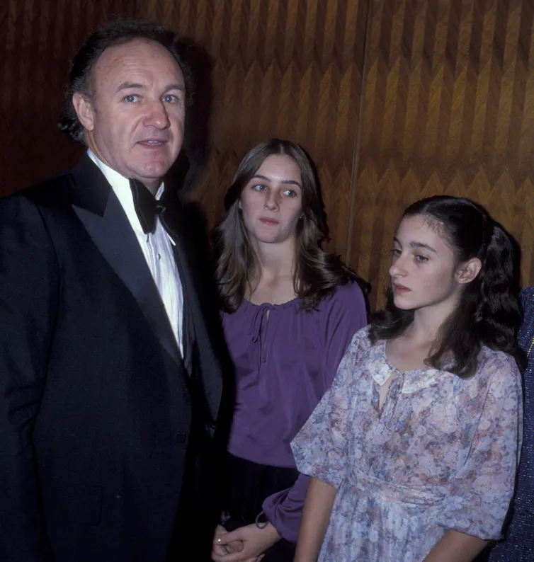 Man in tuxedo and a large bowtie stands next to two teenagers who are looking away.