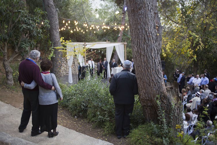 A couple embraces side by side as they observe a small wedding in a wooded area.
