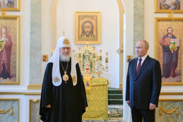 Two men, one in black clerical robes, stand stiffly in an ornate room with gold-framed paintings.