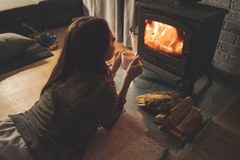 Woman lying down on floor staring at wood stove, holding mug of tea or coffee