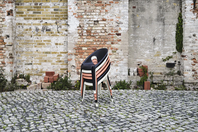 A stack of salmon pink, black and white plastic chairs on a cobbled surface, set against a rustic brick wall
