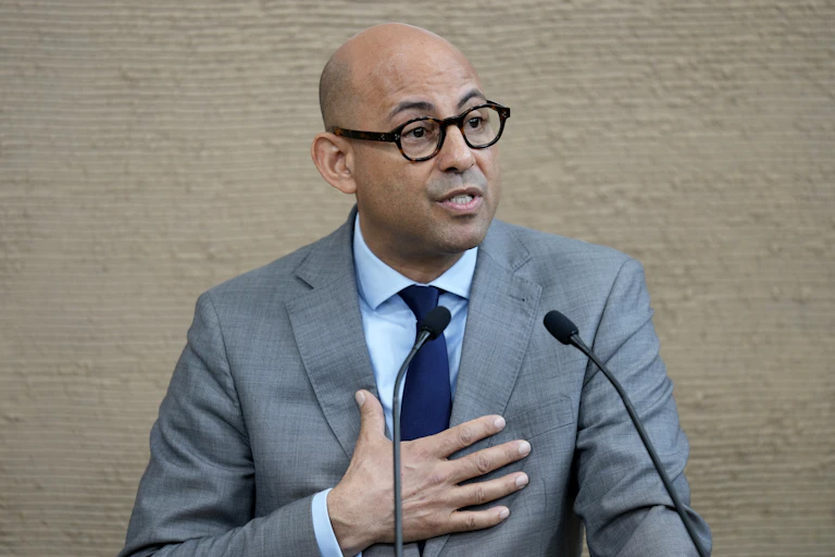 A bald man in glasses and a suit speaks at a podium