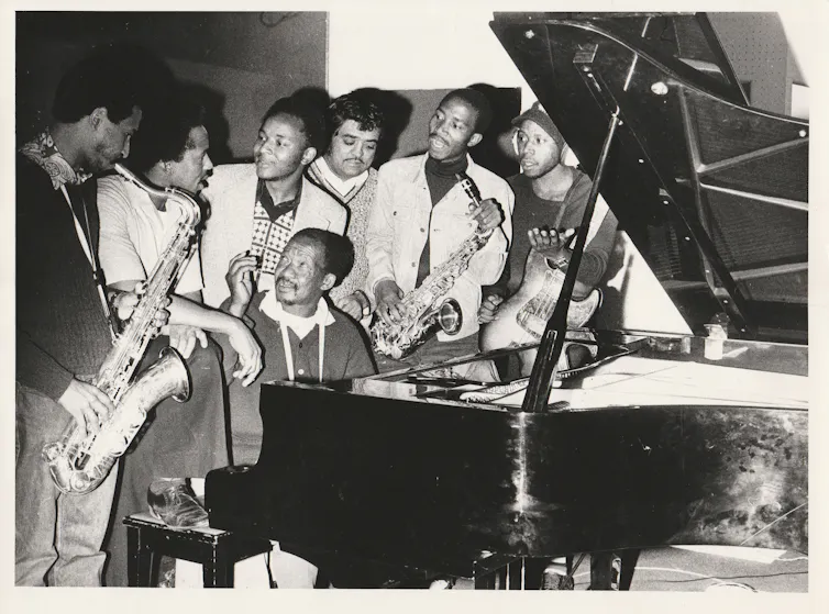 An old black and white photo of young to middle-aged African men, one seated at a paino and surrounded by the others, some holding musical instruments.