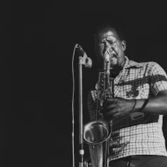 A black and white photo of an African man playing the saxophone at a mic.