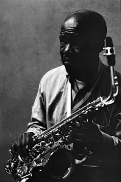 A black and white studio portrait of a balding African man holding a saxophone, his face mostly in shadows.