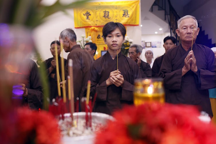 A young man, holding incense sticks, stands with an elderly person while they both fold their hands in prayer at an altar, with several others behind them.