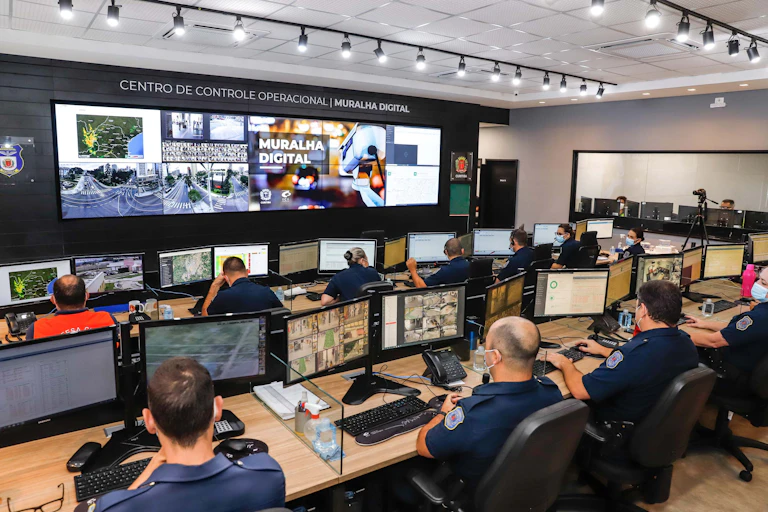 Pessoas sentadas olhando para seus computadores numa bancada e, ao fundo, para um painel repleto de monitores.