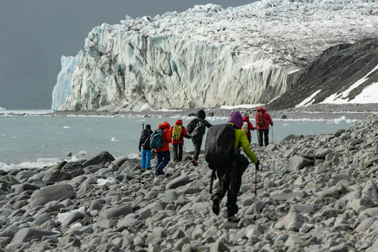 Personas caminando por un terreno pedregoso con el formaciones montañosas y paisajes helados al fondo
