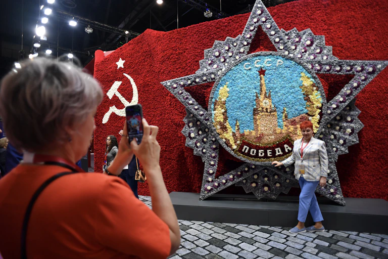 A woman takes a picture of another woman next to a display with a hammer and sickle design