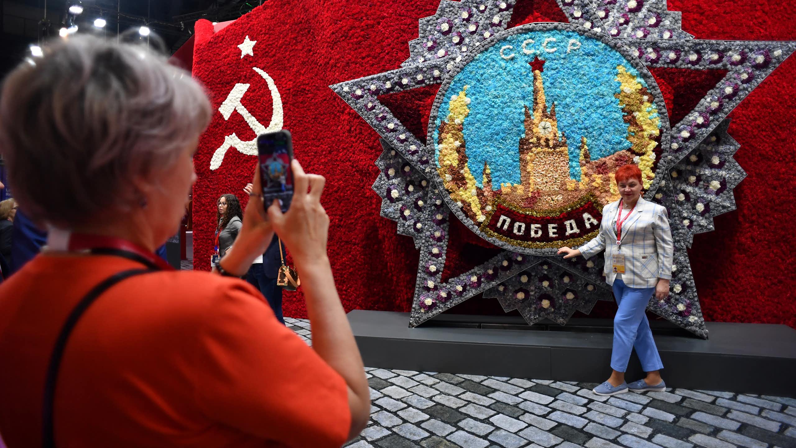 A woman takes a picture of another woman next to a display with a hammer and sickle design