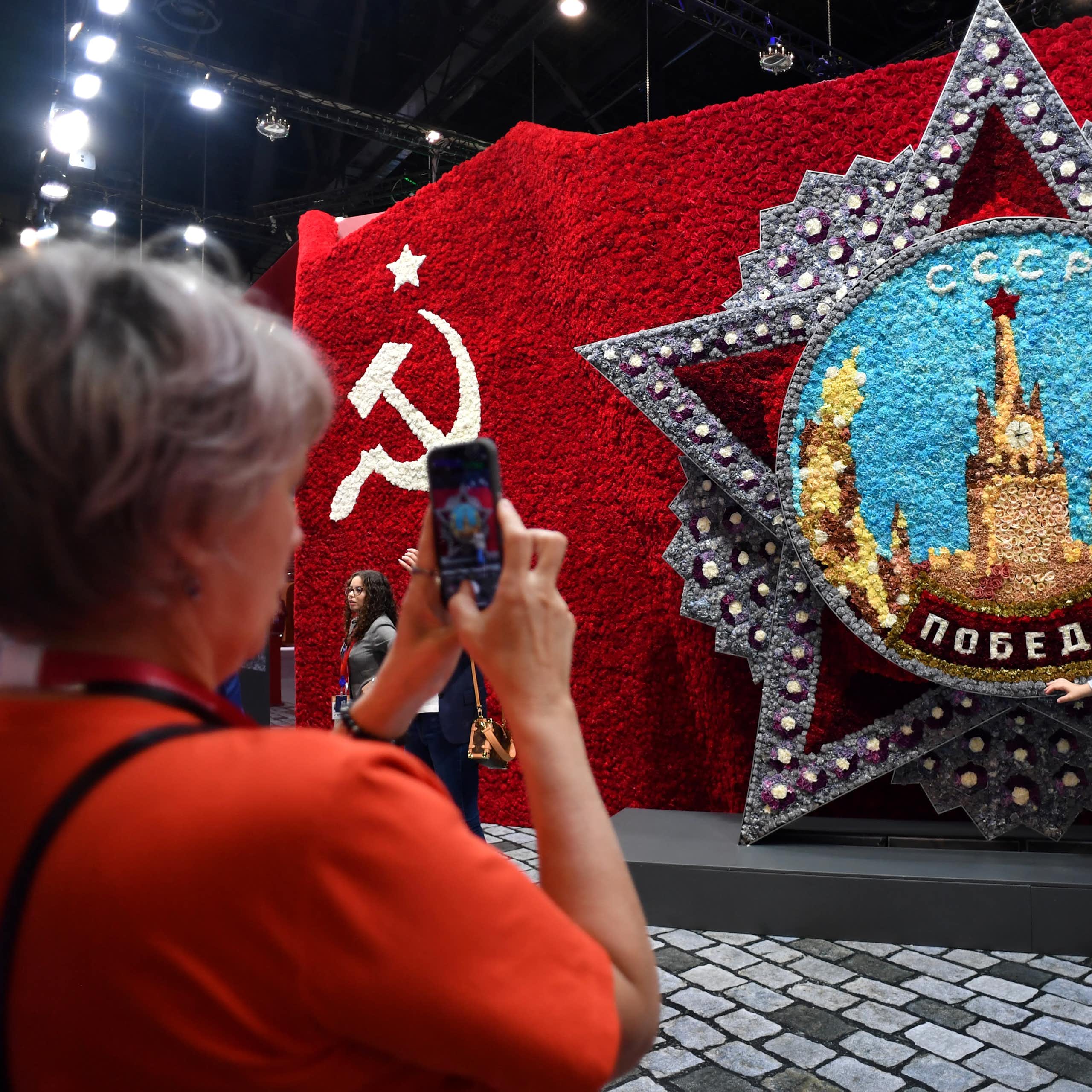 A woman takes a picture of another woman next to a display with a hammer and sickle design