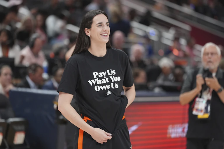 A white woman with long, brown hair wearing a shirt that says 'Pay use what you owe us'