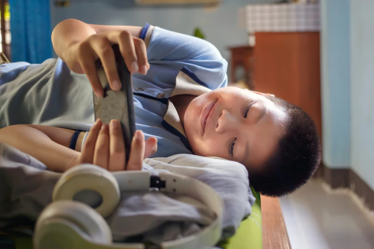 A kid lying sideways on a bed in his room watching a video on his phone and smiling.