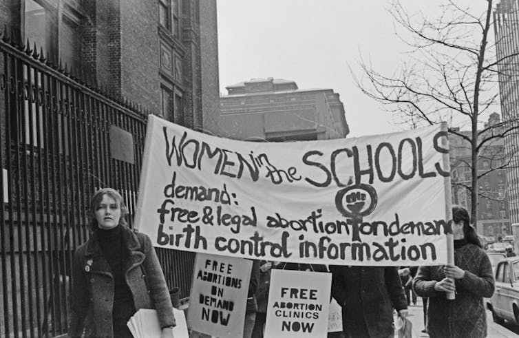 Banner reading 'Women in the schools demand free and legal abortion on demand, birth control information' featuring a raised fist in the circle of the female gender symbol, with placards reading 'Free abortions on demand now'.