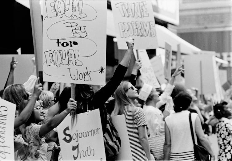 One visible sign reads 'Equal Pay For Equal Work,' while others reference 'Trainee Programs' and Women's and African-American Rights activist Sojouner Truth.