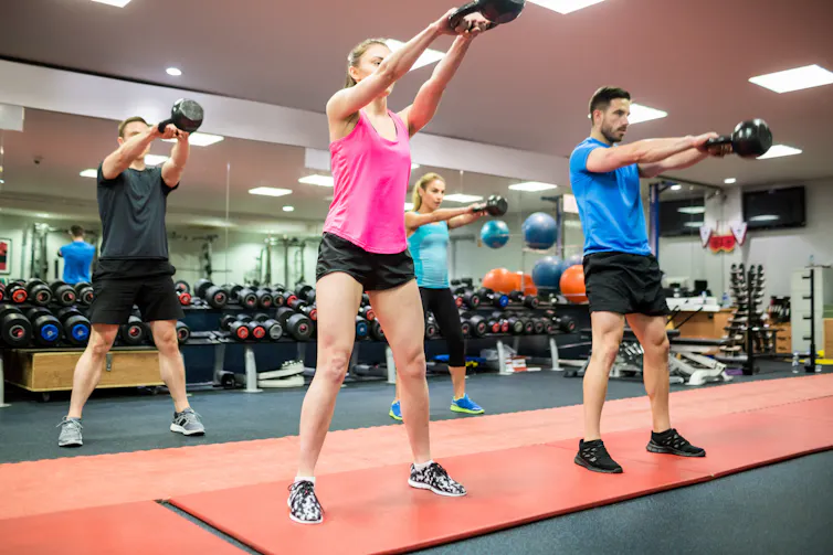A group of young, fit people perform kettlebell swings in a gym.