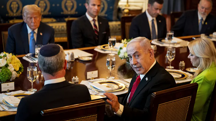 Benjamin Netanyahu turns and looks at the camera while sitting at a table in the White House with US president Donald Trump and others.