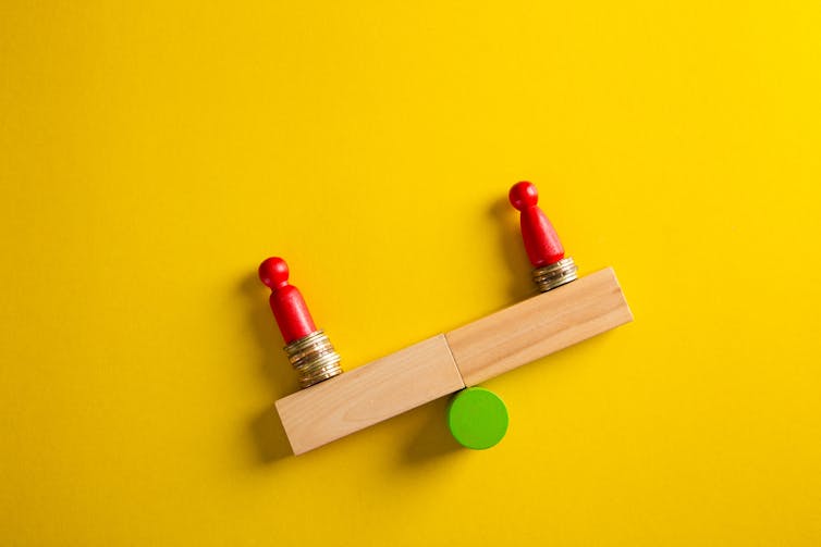 Wooden blocks depicting two people on an uneven seesaw, against a yellow background