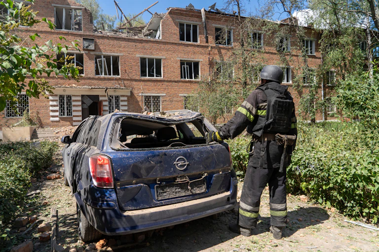 A man in a protective clothing stands next to a damaged car in front of a bombed hospital.
