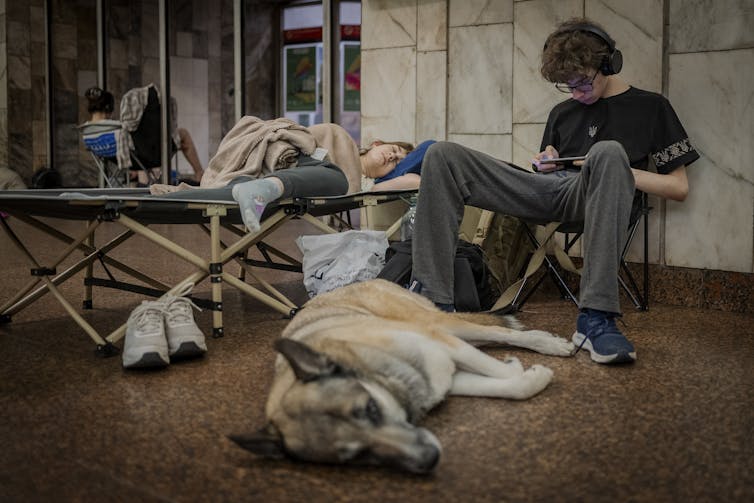 El joven está sentado en la estación de metro con su perro durmiendo sobre sus pies.