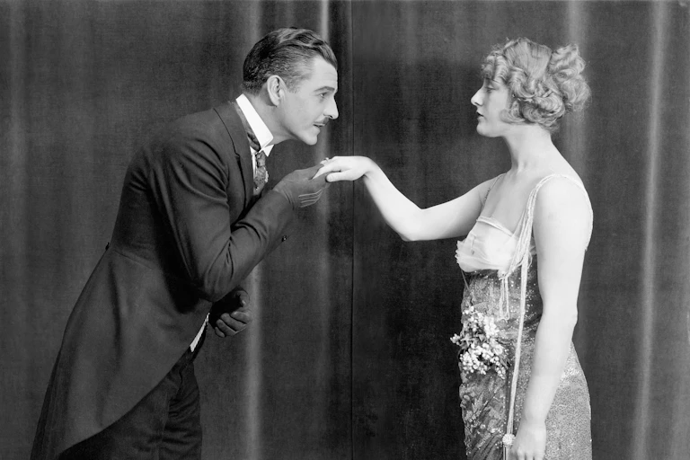 Una fotografía en blanco y negro de un hombre vestido de frac besando la mano de una mujer.