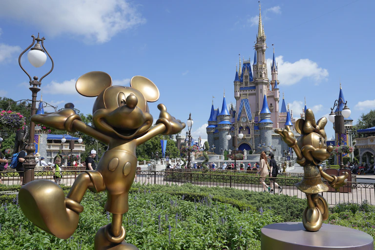 A gold statue of a cartoon mouse next to shining castle against a blue sky.