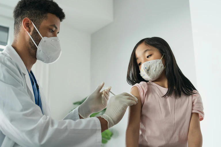 A girl in a face mask getting an injection from a health-care professional in a white coat and face mask