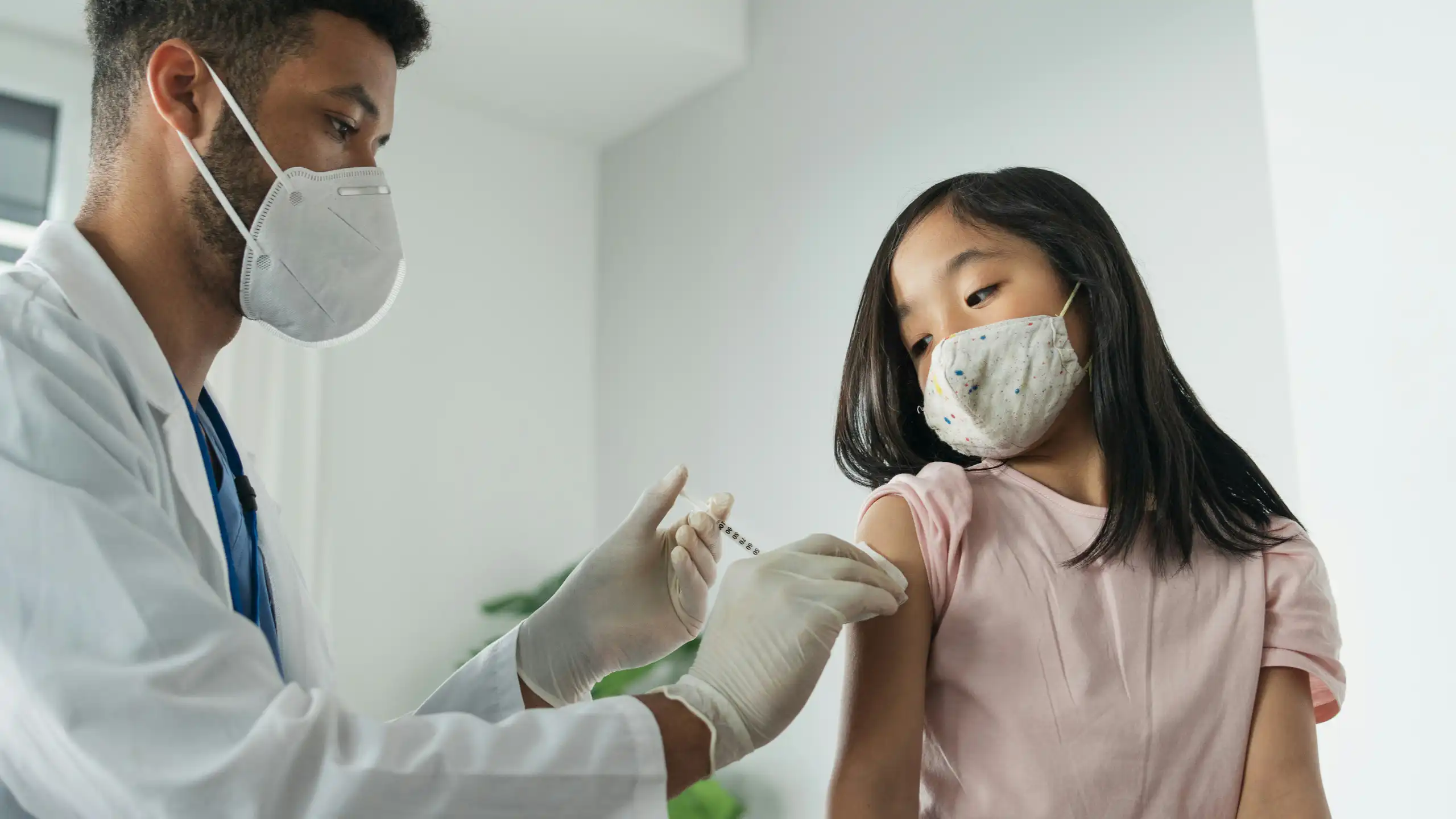 A girl in a face mask getting an injection from a health-care professional in a white coat and face mask