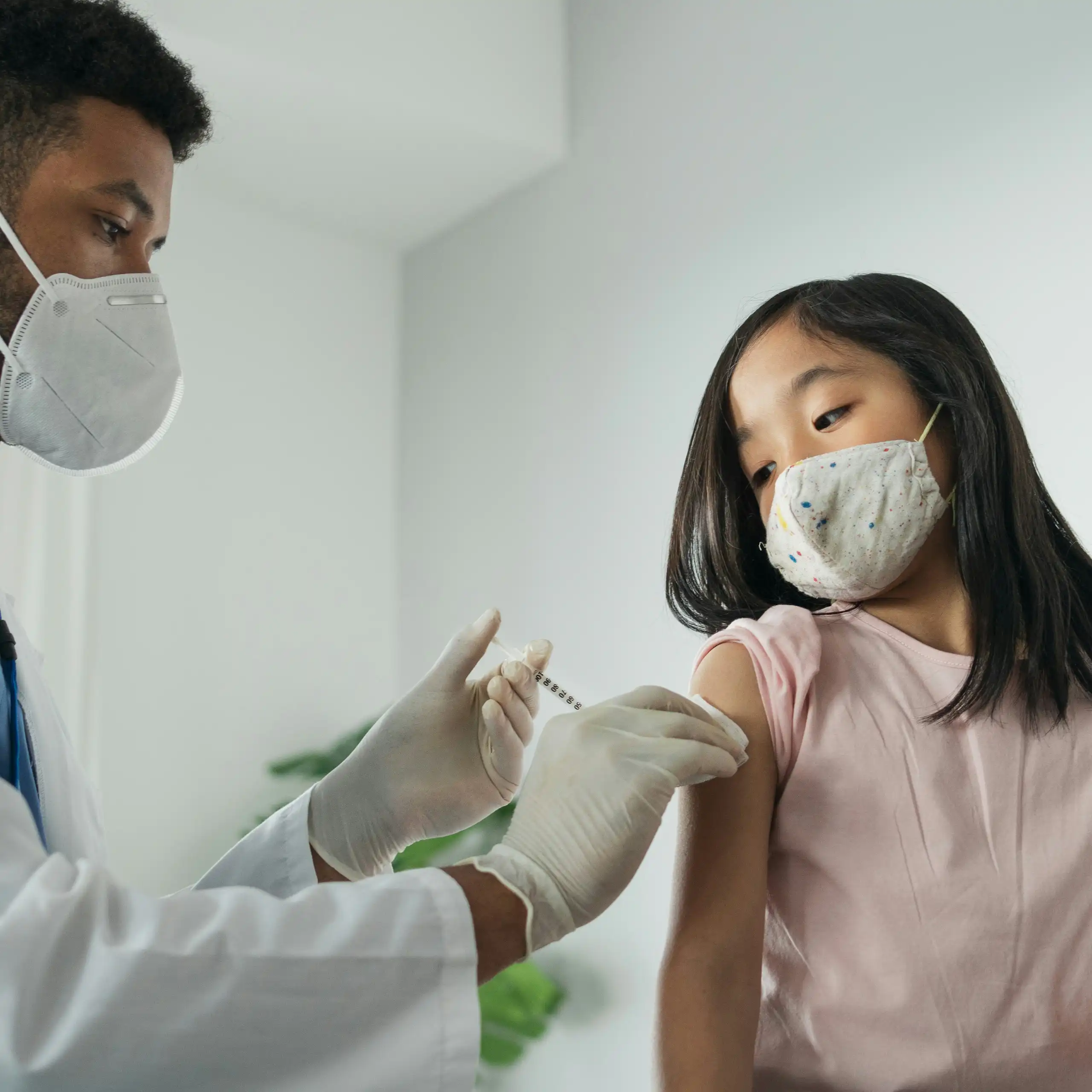 A girl in a face mask getting an injection from a health-care professional in a white coat and face mask
