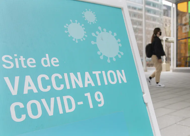 A blue sandwich board sign reading 'Site de vaccination COVID-19' with a person out of focus in the background