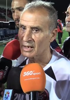 Un homme chauve, édenté, parle dans plusieurs microphones sur un terrain de football.