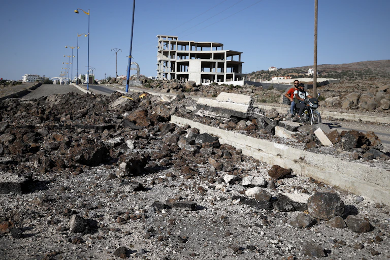 Militiamen on a motorcycle ride along a road surrounded by rubble with a solitary empty building in the background.