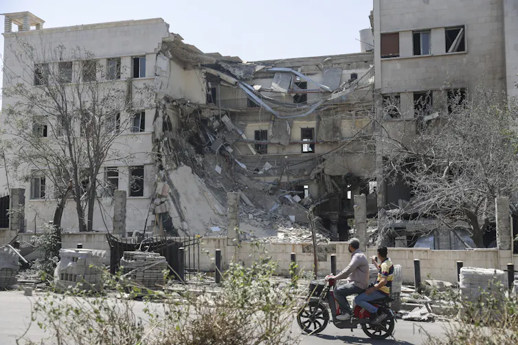 People on motorbikes and on foot walk past a damaged government building.