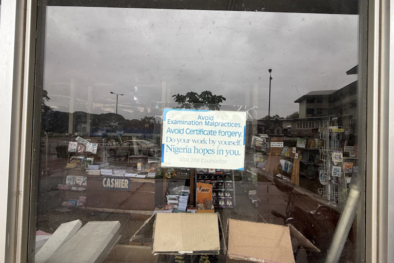 A shop window with books sprawling in the distance and some on disolay in front. A sign pasted to the window reads, "Avoid examination malpractices. Nigeria hopes in you."