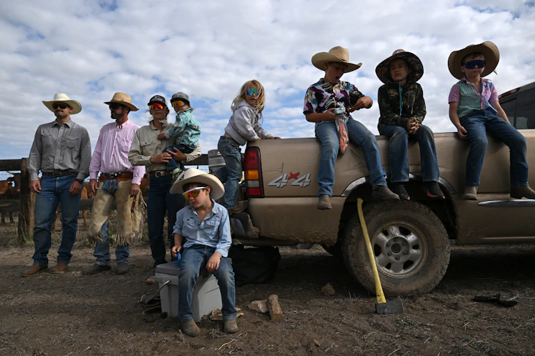 A group of adults and children, many wearing cowboy hats, gather near a pickup truck.